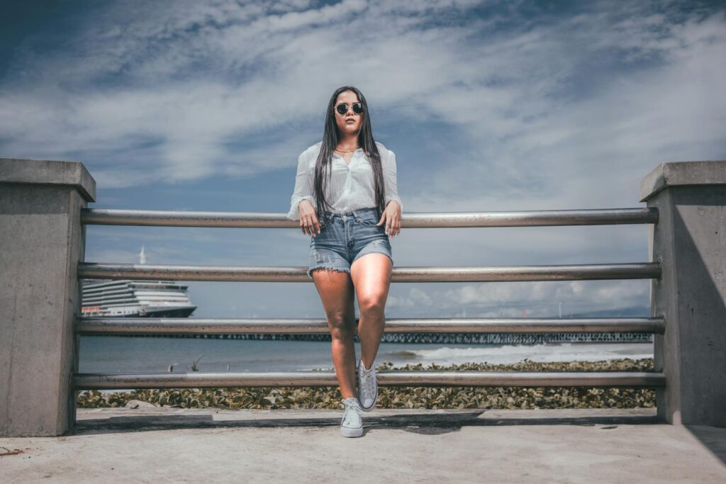 Woman in Shorts and Sunglasses Standing on a Pier on the Background of Sea and Ship